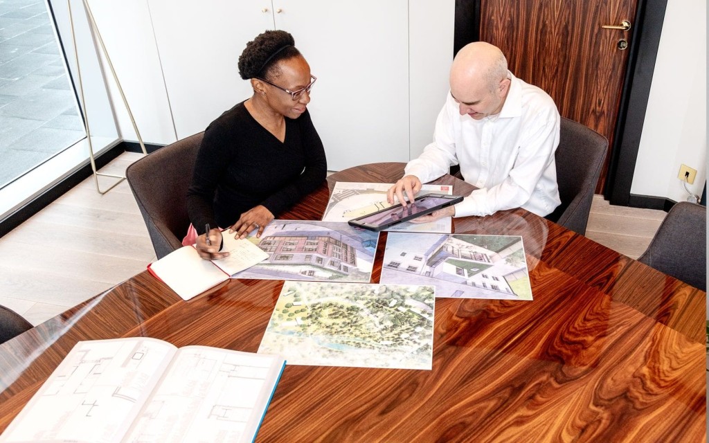 Two architects, a woman and a man, engaged in discussion over house plans and digital tablets at a wooden table, with architectural drawings and designs spread out in front of them.