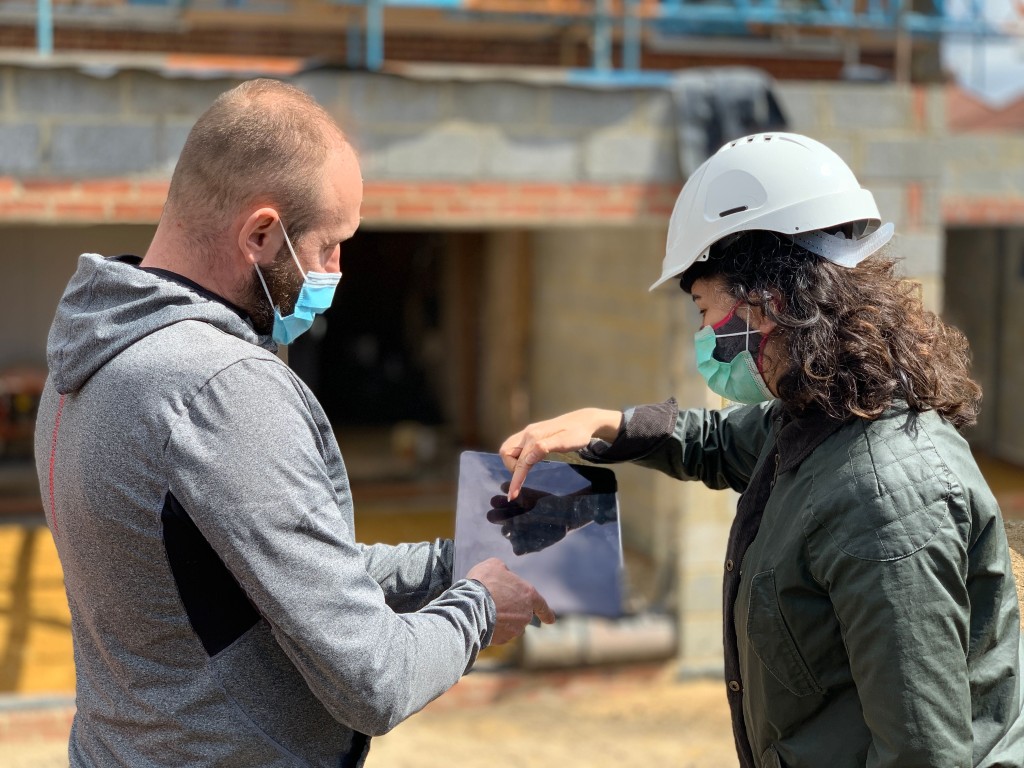 Two construction professionals wearing safety helmets and face masks discussing over a tablet at a building site with a house under construction in the background.