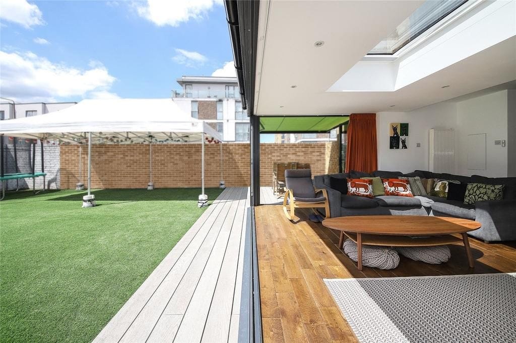 Contemporary outdoor living space with synthetic lawn and gazebo next to a cosy interior with wooden flooring, large sofa, and skylight.