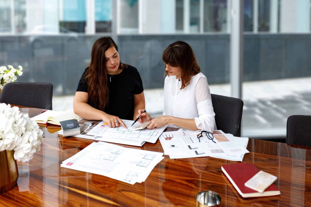 Two female architects collaborating over house blueprints on a polished wooden table in a modern office setting with glass walls.