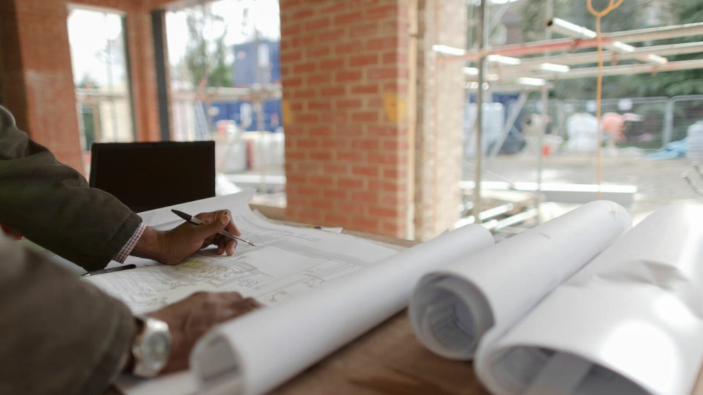 Focused engineer working on architectural blueprints at a construction site, with a clear view of the ongoing work in the background, illustrating active project management.