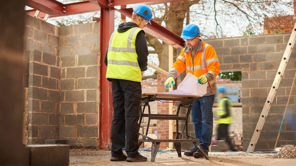 Two construction workers in safety gear and hard hats consulting building plans on a workbench at a construction site with cinder block walls and steel beams in the background.