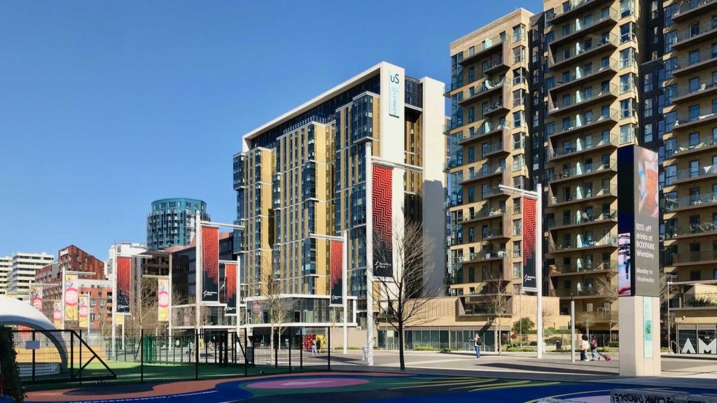 Modern urban landscape showcasing a mix of residential high-rise buildings with gold and glass facades, a round blue-glass office building, and red patterned banners along a pedestrian plaza, with a foreground of a colorful playground and green sports court, under a clear blue sky.