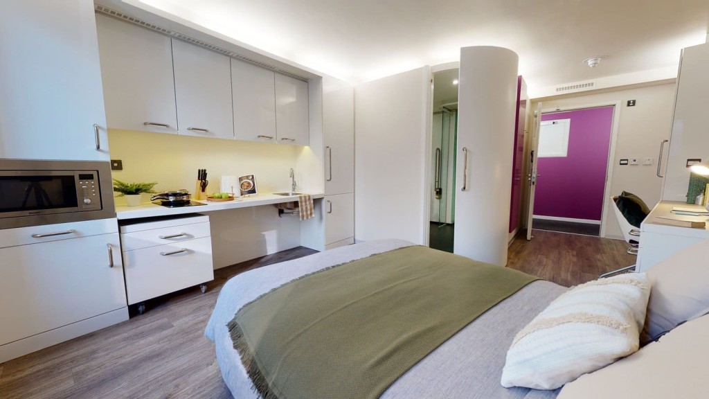 Compact and modern student studio apartment with a streamlined kitchenette featuring stainless steel appliances, white cabinetry, and a subtle yellow backsplash, alongside a cosy bed with olive green throw, leading to a bright hallway with a vibrant magenta door.