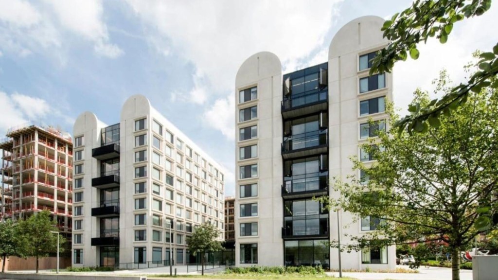 Contemporary architecture of twin residential buildings with distinctive rounded tops and black balconies, complemented by a construction site in the background, greenery in the foreground, on a sunny day with partial cloud cover.