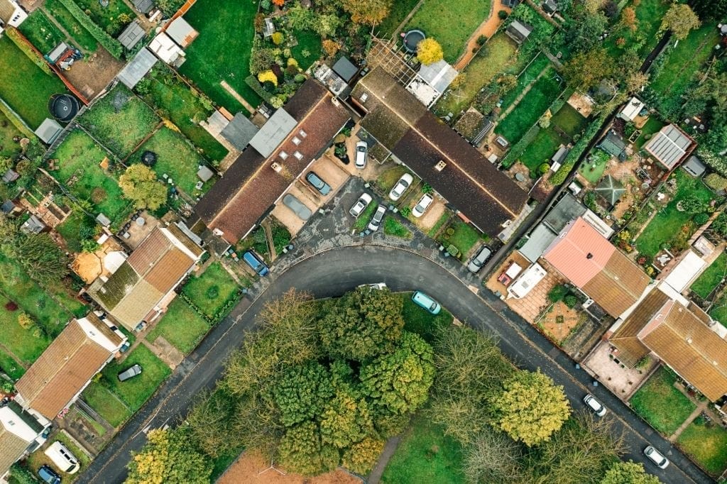 Aerial view of a residential area with a curved street, showcasing a variety of houses with gardens and vehicles parked alongside, surrounded by greenery, exemplifying suburban neighborhood planning.