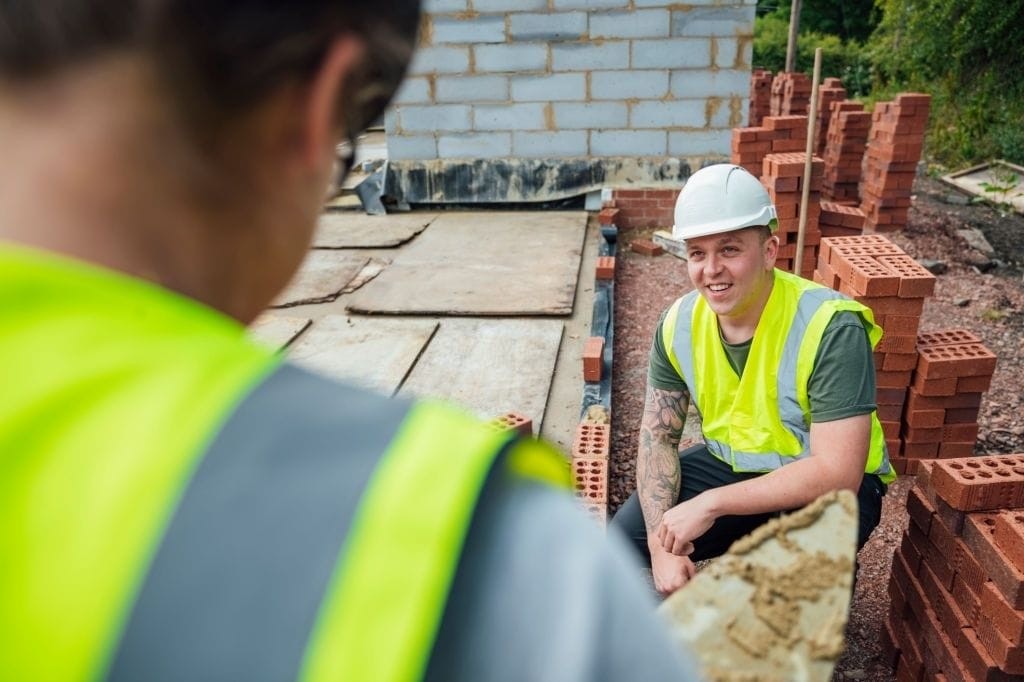Smiling construction worker in high visibility vest and safety helmet sitting on a brick wall at a construction site with another worker in the foreground, showcasing teamwork and safety in the building industry.