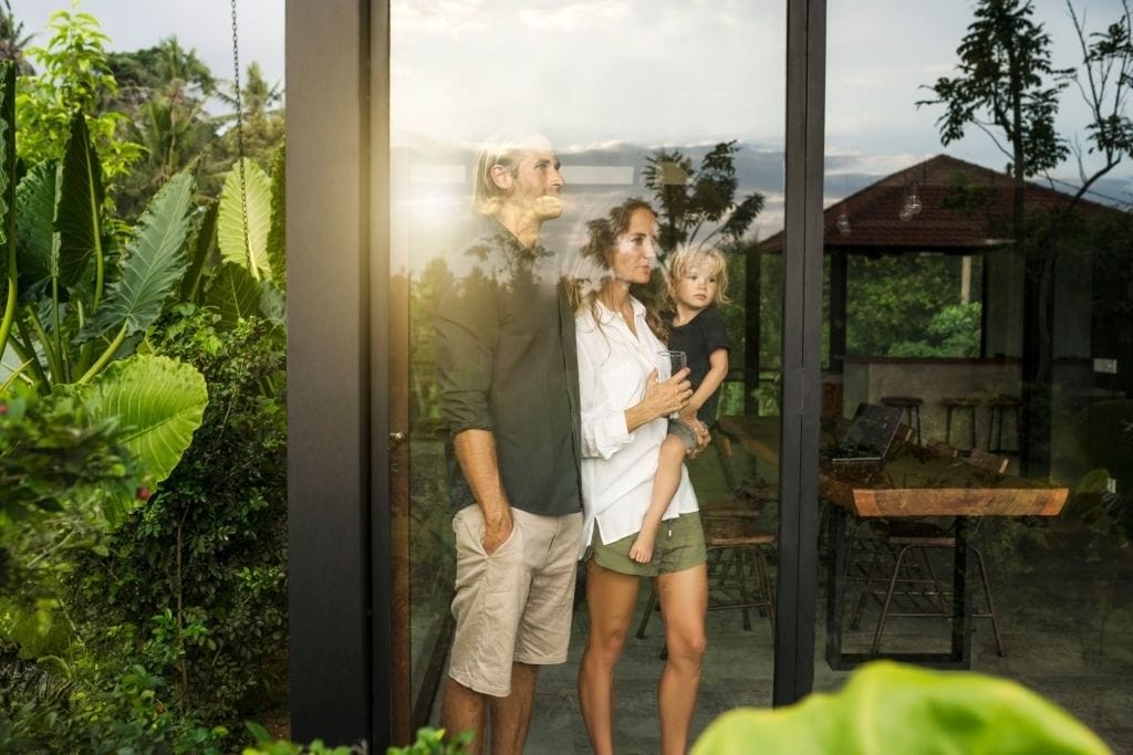 Family embracing sustainable living, standing inside a modern eco-friendly house with large glass windows overlooking a lush tropical garden, reflecting a connection with nature.