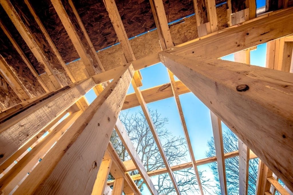 Upward view of a wooden roof structure under construction with beams and rafters illuminated by sunlight, showcasing sustainable building materials and design in home renovation.