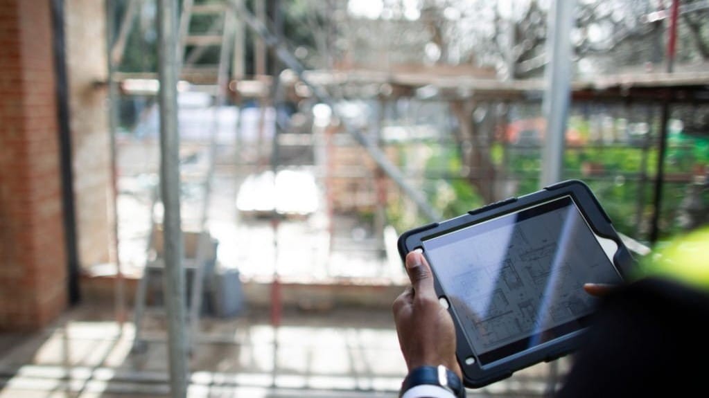 Professional reviewing architectural plans on a digital tablet at a construction site with visible scaffolding in the blurred background.