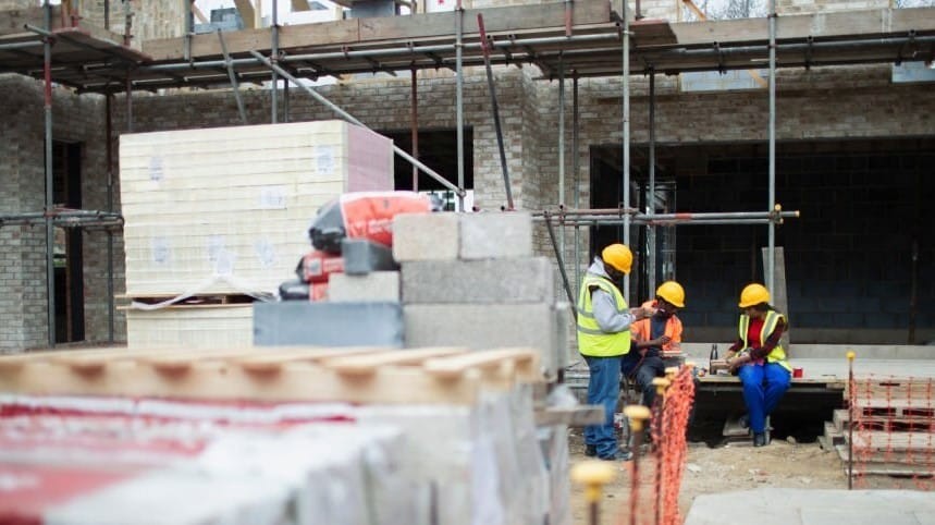 Construction workers with hard hats engaging in a discussion at a building site with scaffolding and construction materials in the foreground.