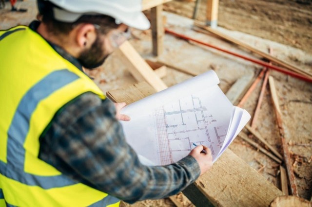 Image cover for the article: Professional architect reviewing blueprints in a spacious room under renovation with exposed wooden beams, natural light, and construction materials.