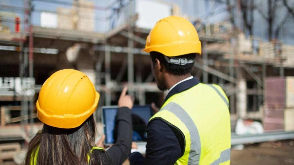 Two construction engineers in hard hats and reflective vests discussing over a tablet at a construction site with scaffolding in the background.