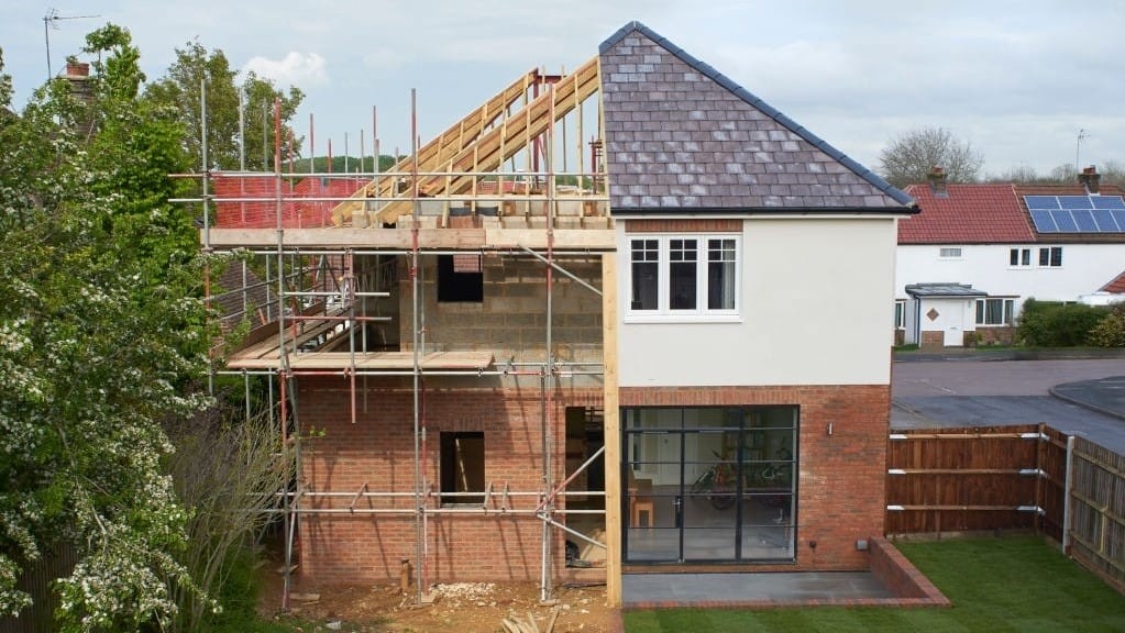 Residential house extension under construction with scaffolding, showing a mix of finished areas and exposed framework against a backdrop of a suburban neighbourhood.
