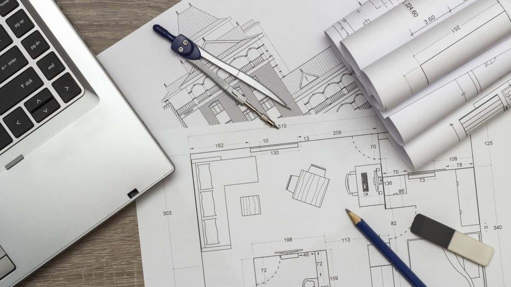 Architectural planning with detailed house blueprints, a laptop, drafting compass, and pencil on a wooden desk, showcasing the design phase of home construction.