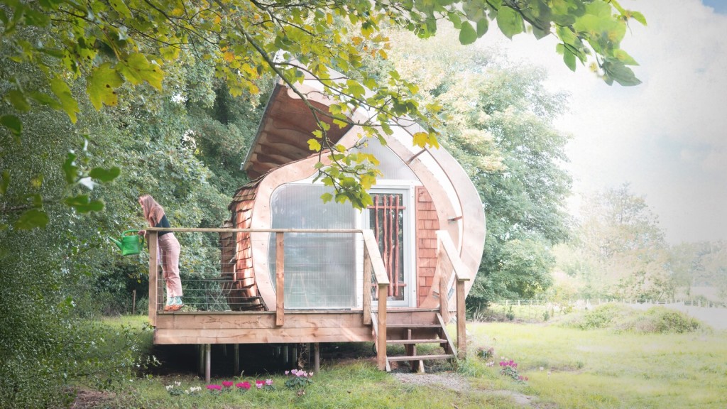 A nature-inspired tiny house with a curved, timber-clad exterior and a small raised deck, surrounded by greenery, with a person watering plants in the countryside.