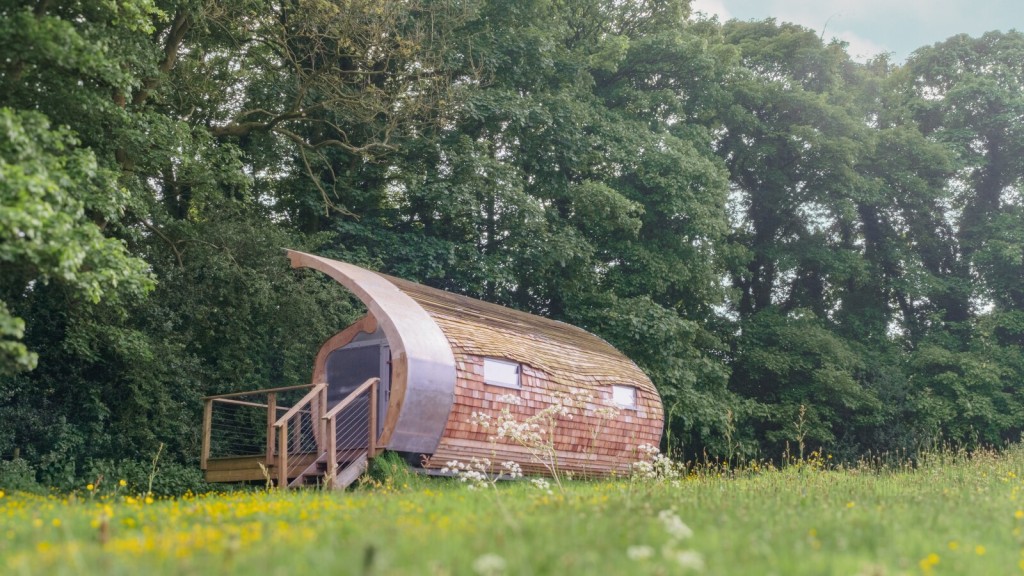 A curved-roof tiny house with timber cladding and a raised wooden deck, surrounded by wildflowers and lush green trees in a peaceful countryside setting.