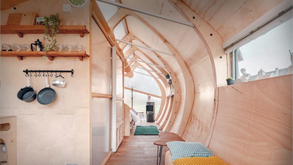 Interior of a curved-roof tiny house featuring wooden walls, open shelving with jars and plants, hanging cookware, and a cozy living area leading to a bed and a small wood stove.