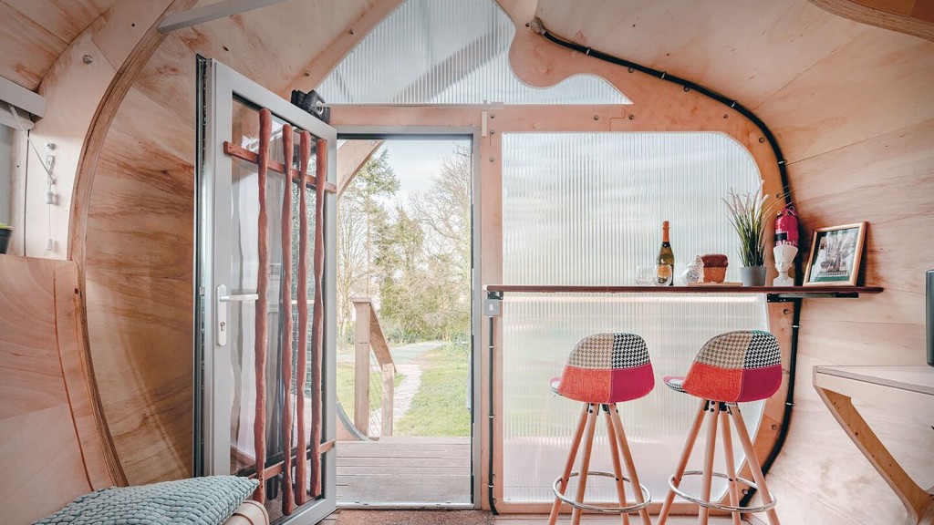 Interior of a tiny house with wooden walls and a curved design, featuring a bar table with two colourful stools, a door with natural wooden details, and large translucent panels letting in soft natural light.