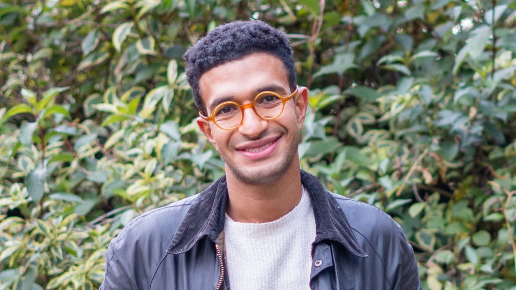 Peter Markos, a tiny home architect, smiling outdoors, wearing a leather jacket, a white sweater, and round orange glasses, with a background of lush green foliage.