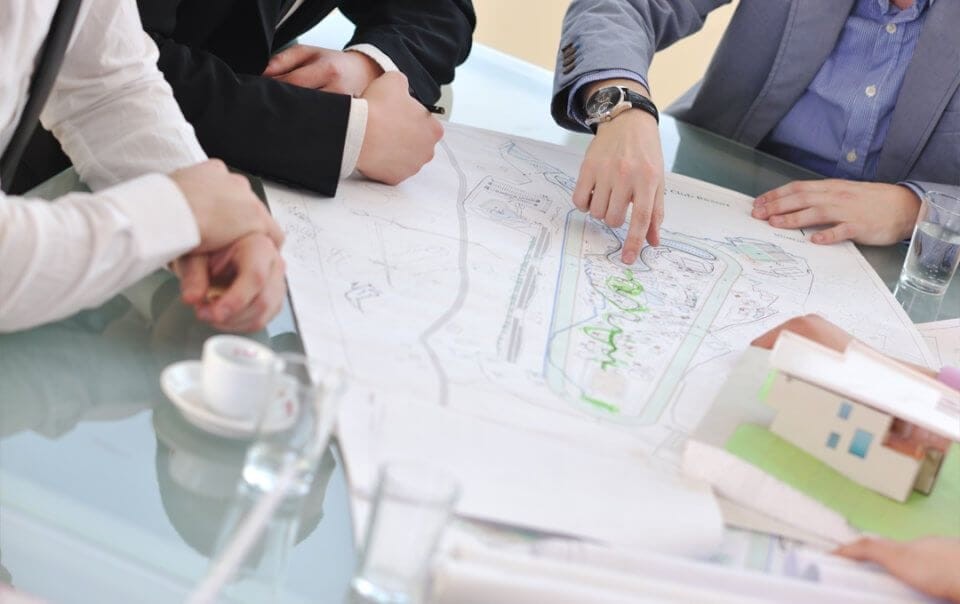 London architects discussing a residential development plan, with a focus on a blueprint and a model house, highlighting architectural planning and design collaboration.