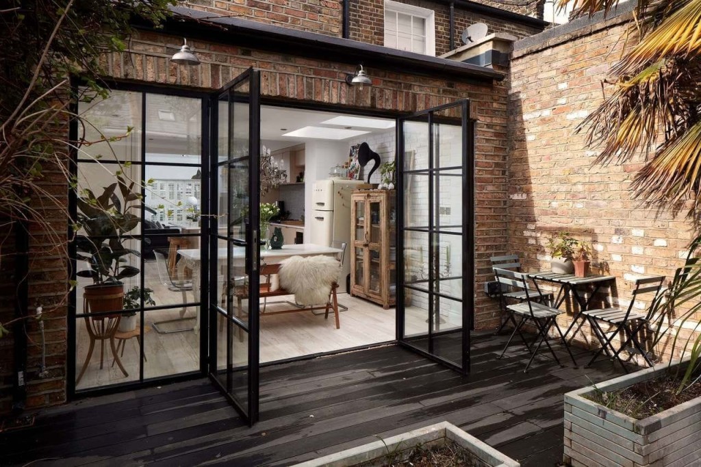Chic urban patio with black framed glass doors opening to a cosy, stylish interior with exposed brick walls, exemplifying modern London home design trends.