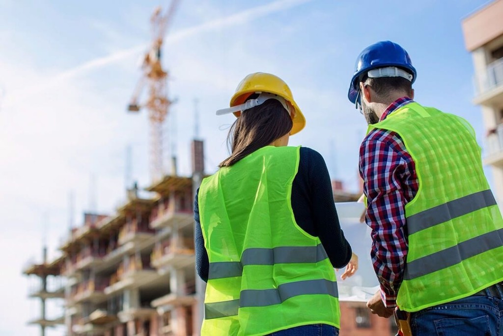 Back view of a male and female construction professionals in safety gear observing an active building site, highlighting the collaboration and oversight in construction management.