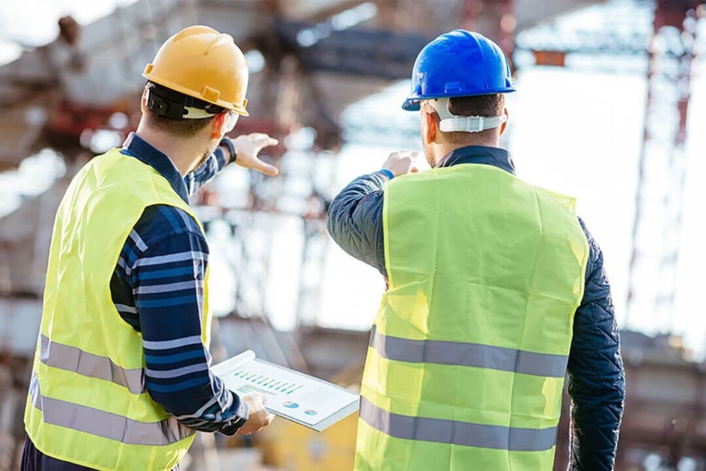 Two construction supervisors in high-visibility vests and safety helmets actively discussing over a chart, with the blurred background of a busy construction site.