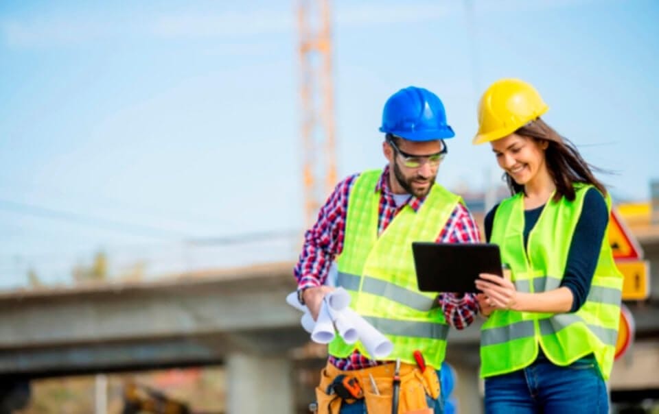 Smiling male and female construction engineers in safety vests and hard hats discussing project details on a digital tablet at a construction site, with blueprints in hand and a crane in the background.