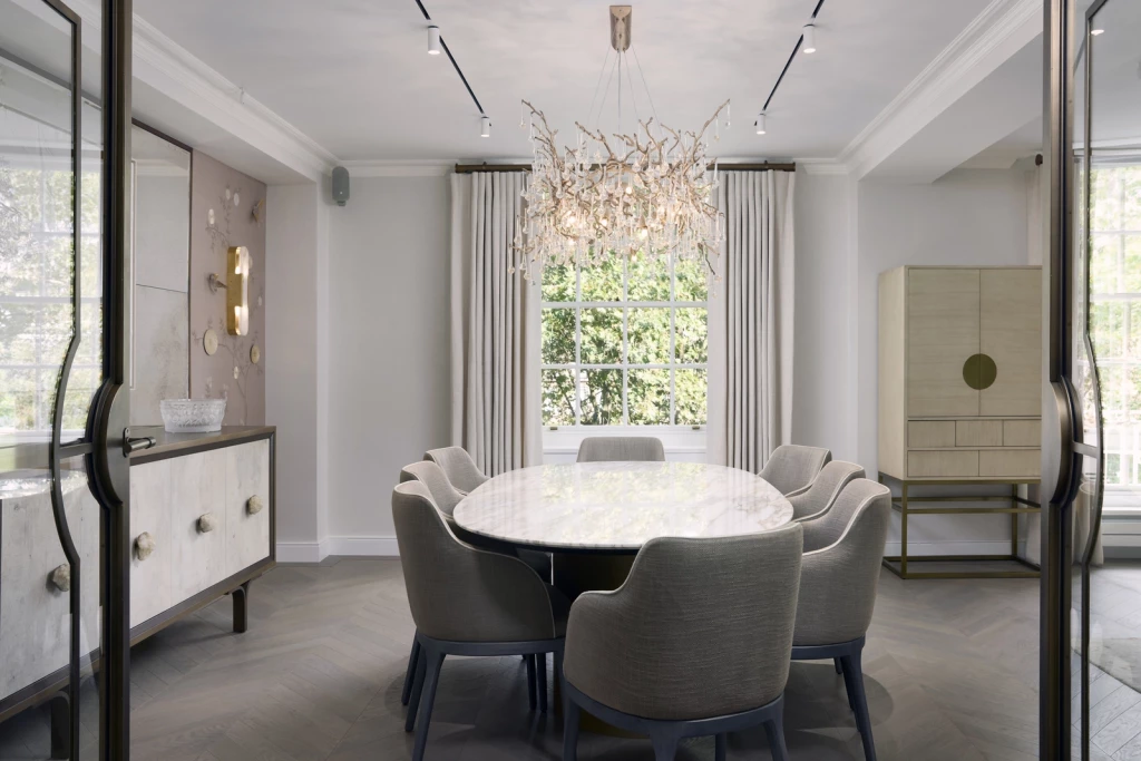 Contemporary dining suite featuring an elliptical marble table and eight upholstered tub chairs beneath a sculptural branch chandelier, with herringbone oak floor, bespoke credenza with sculptural pulls, brass-detailed armoire and linear track lighting, daylight moderated by full-height curtains over a multi-pane window.