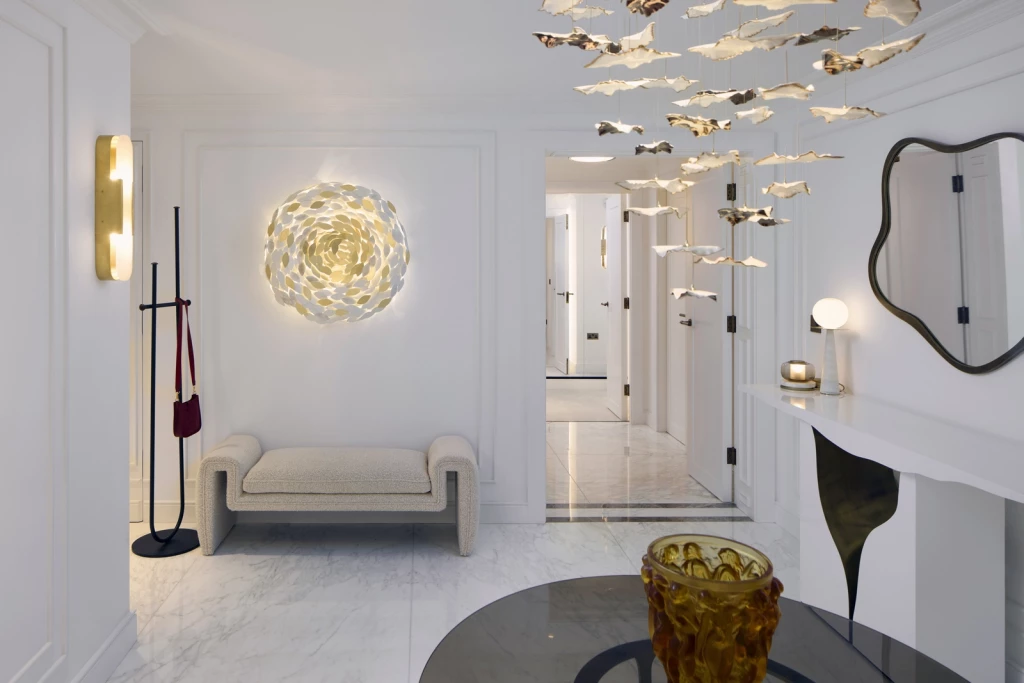 Refined hallway interior with sculptural hanging light installation, marble flooring, curved bench, illuminated wall art resembling layered petals, asymmetric mirror, and minimalist console with spherical lamp and candle.