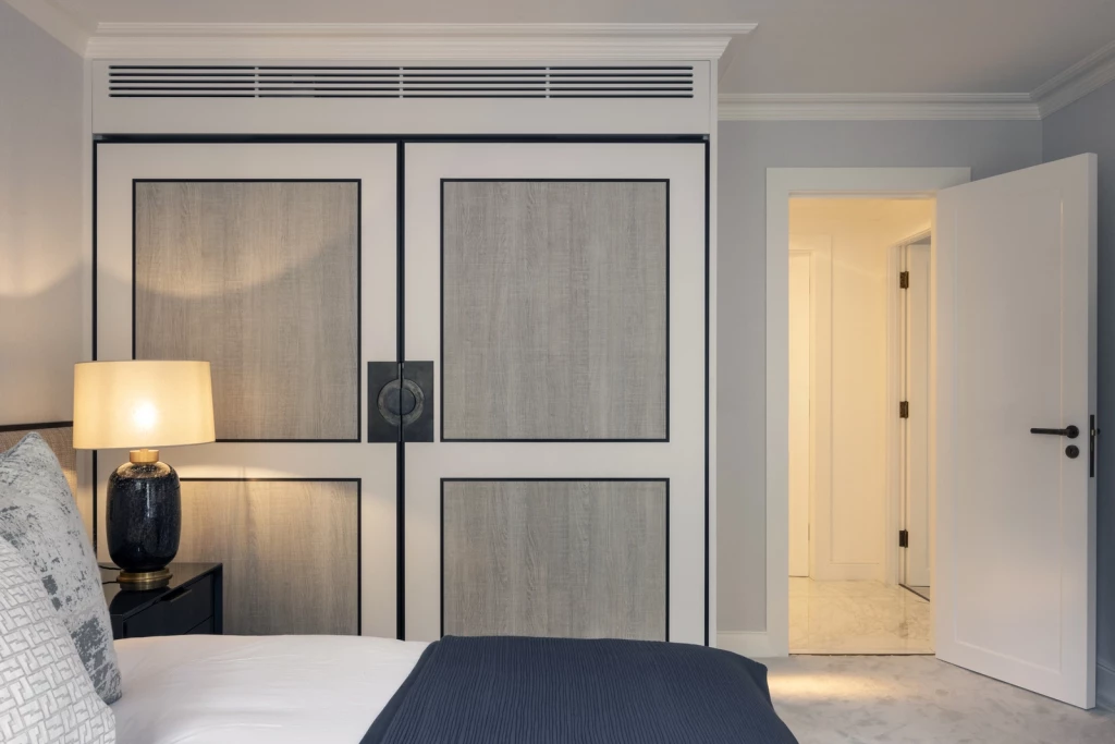 Bedroom interior featuring custom wardrobe with wood-grain panels framed in black, central circular bronze handles and overhead ventilation grille; bedside table with dark ceramic lamp, soft grey textiles and open doorway leading to marble-floored corridor.