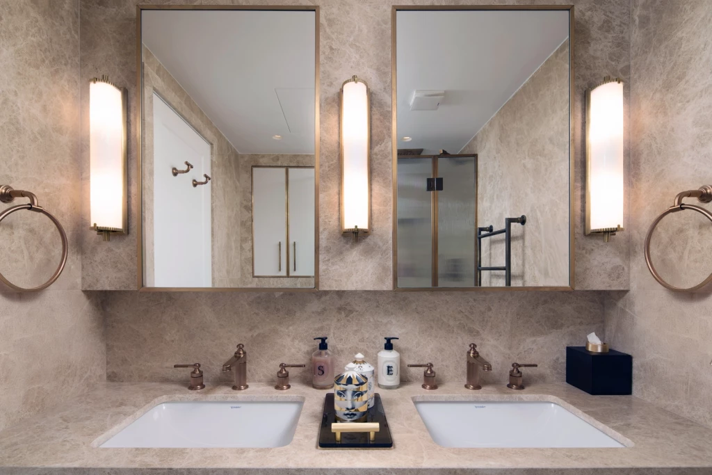Bathroom interior with double vanity in beige marble, two integrated basins, bronze fixtures, twin mirrors with vertical wall lights, and recessed cabinetry reflected in the mirrors; accessories include decorative jars and soap dispensers.