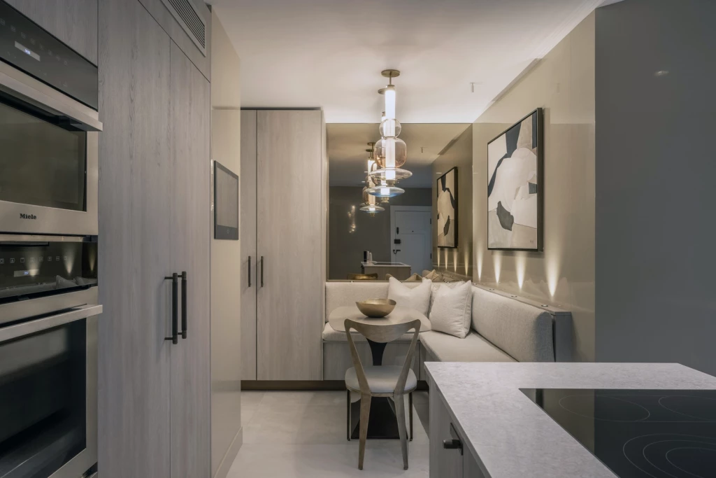 Compact contemporary kitchen with integrated appliances, full-height cabinetry in light wood tones, stone worktops, and a built-in dining nook with upholstered banquette, sculptural dining chair and small round table, accented by pendant lighting and abstract artwork.