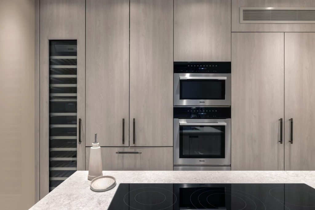 Minimalist kitchen wall with full-height light wood cabinetry, integrated wine fridge, built-in Miele oven and microwave, concealed storage, black handles and overhead ventilation grille, complemented by a stone island worktop with induction hob.