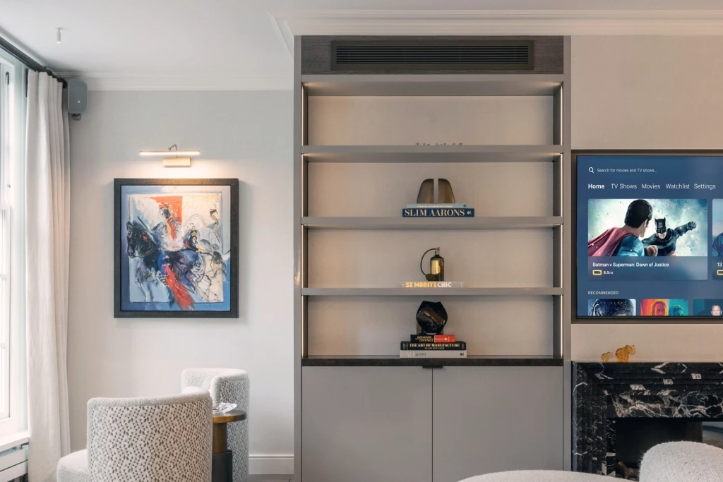 Living space with integrated shelving unit styled with books and decorative objects, adjacent wall-mounted TV above a black marble fireplace, artwork with picture light on the opposite wall, and upholstered armchairs with side table by the window.