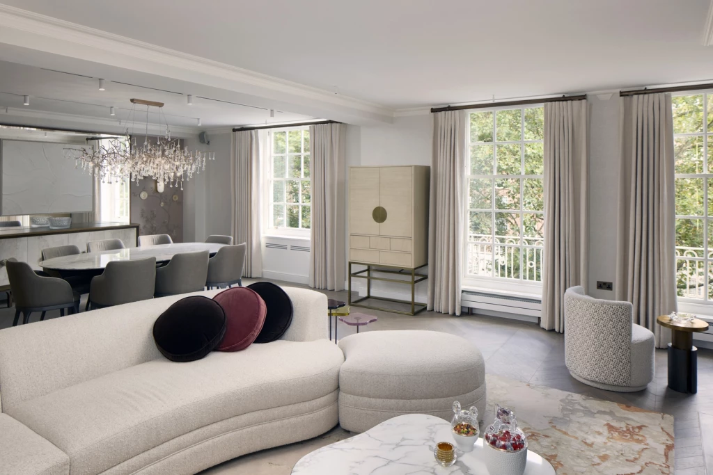Open-plan living and dining interior featuring a curved cream sofa with round ottoman and marble coffee table in the foreground; to the rear, an elliptical marble dining table with eight upholstered chairs under a sculptural branch chandelier, bespoke sideboard and brass-detailed cabinet, herringbone oak floor, and full-height curtains framing multi-pane windows.