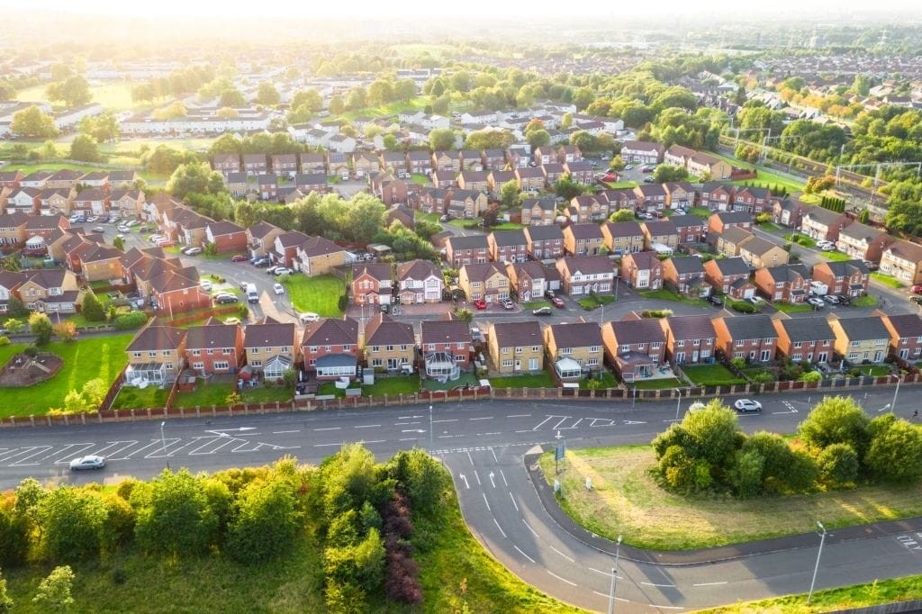 Aerial view of a British suburban neighbourhood at sunset with rows of houses, lush green gardens, and empty roads indicating a quiet residential area.