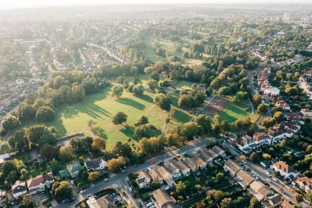 Drone shot of a leafy residential area in the UK during golden hour, featuring community green spaces and a mix of detached homes and tree-lined streets, highlighting the balance of urban and natural elements in suburban planning.