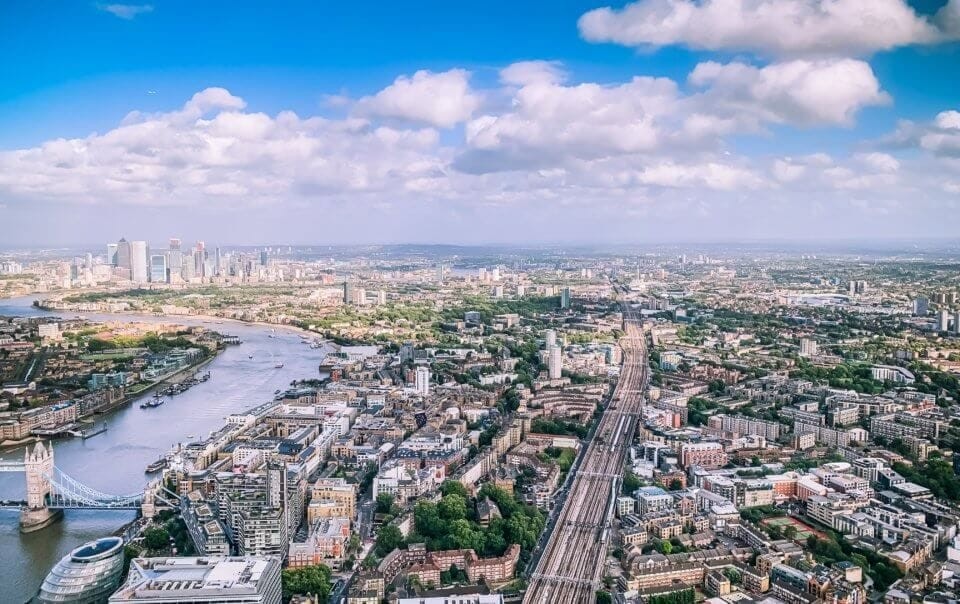 Aerial view of London showcasing the River Thames, Tower Bridge, and surrounding urban landscape under a partly cloudy sky.