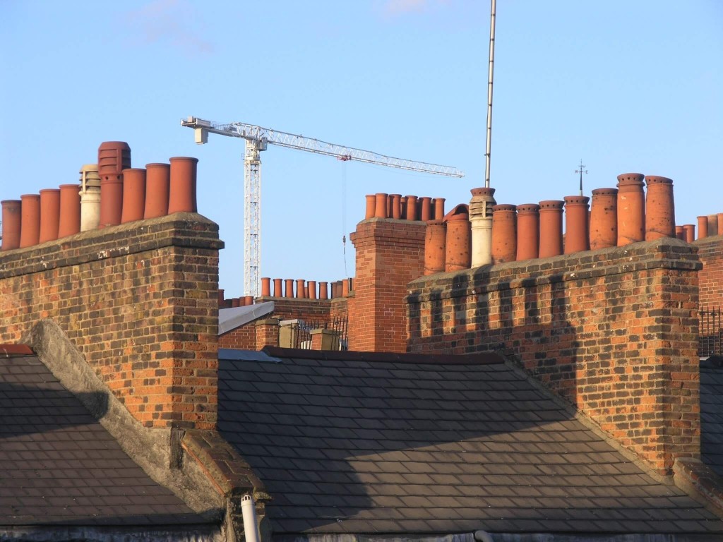 Close-up of terracotta chimney pots on brick rooftops with a construction crane in the background against a clear blue sky, symbolising ongoing urban development and architectural heritage.