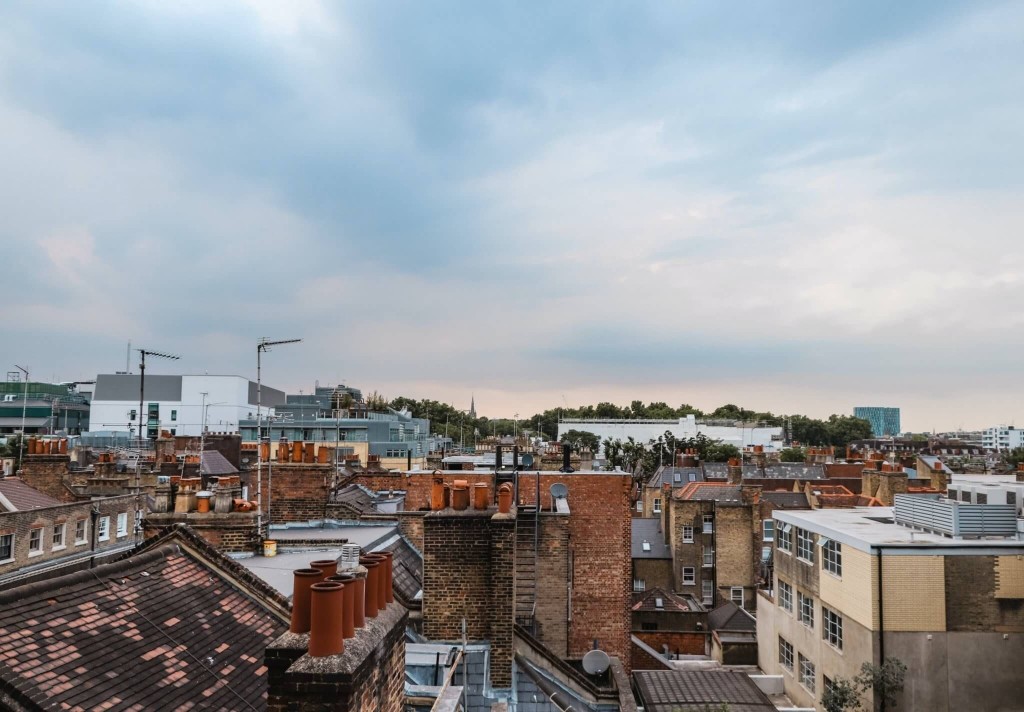 Rooftop view of a diverse urban landscape at dusk, highlighting the architectural variety with a mix of old brick chimneys and modern flat rooftops against a soft cloudy sky.