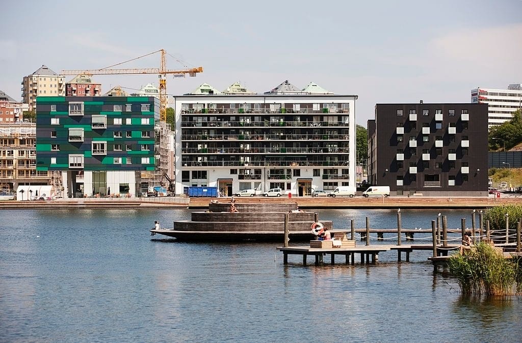Urban waterfront development showcasing a blend of modern architecture with green and white facades, and a distinctive black building with unique window patterns, next to a tranquil city lake with a wooden pier and relaxing residents.
