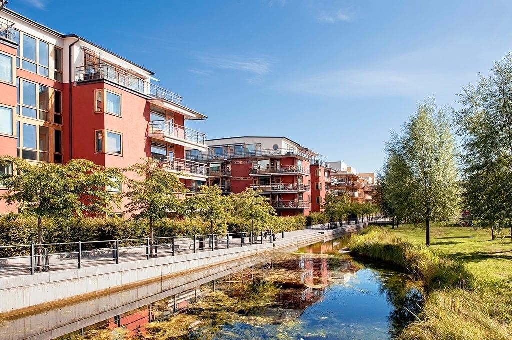 Scenic view of sustainable urban housing along a landscaped waterway, featuring red and beige modern apartment buildings with lush greenery and pedestrian walkways under a clear blue sky.