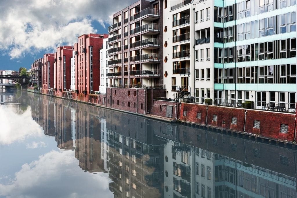 Reflections of modern apartment buildings along an urban canal, showcasing contemporary residential architecture with a mix of red brick and white facades under a clear sky.