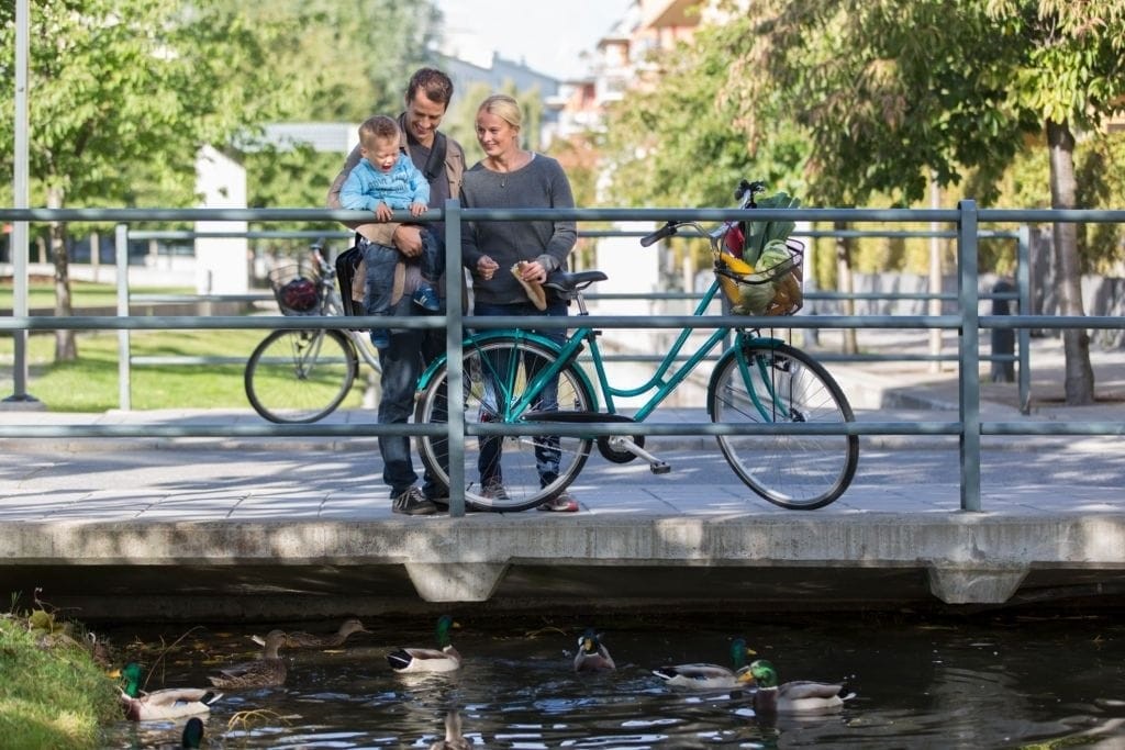 Family enjoying a sunny day in an urban park by a canal with ducks, showcasing a couple with their child and a bicycle, reflecting sustainable city living and active family lifestyle in urban design.