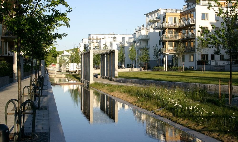 Tranquil urban canal lined with modern residential buildings, lush green landscaping, and a pedestrian pathway, reflecting a harmonious blend of nature and contemporary architecture in a master-planned community.