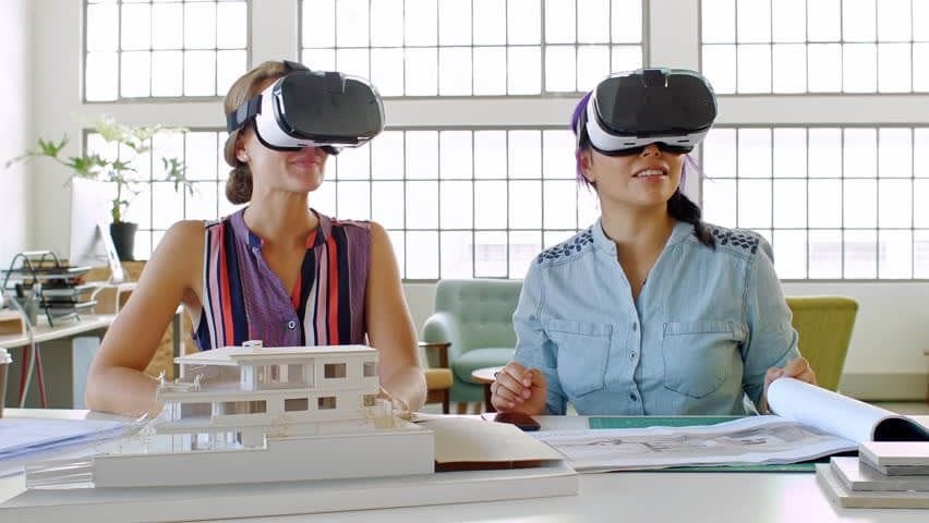 Two female architects wearing virtual reality headsets examining architectural models and plans in a bright office environment, highlighting the use of VR in modern design processes.