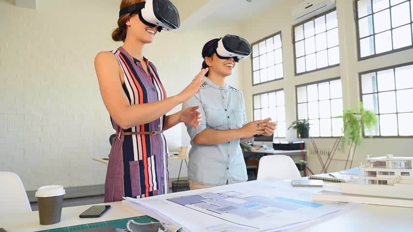 Two smiling female professionals engaging with virtual reality headsets in a well-lit architectural office, interacting with a 3D building plan on a table, illustrating collaborative VR technology in architecture.