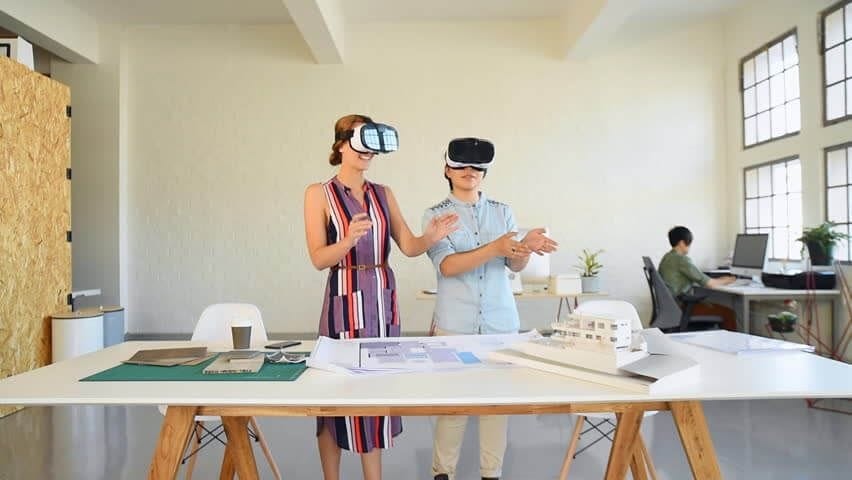 Two architects using virtual reality headsets in a spacious design studio, with a scale model of a building and architectural drawings on the table, demonstrating the integration of VR in architectural visualisation.